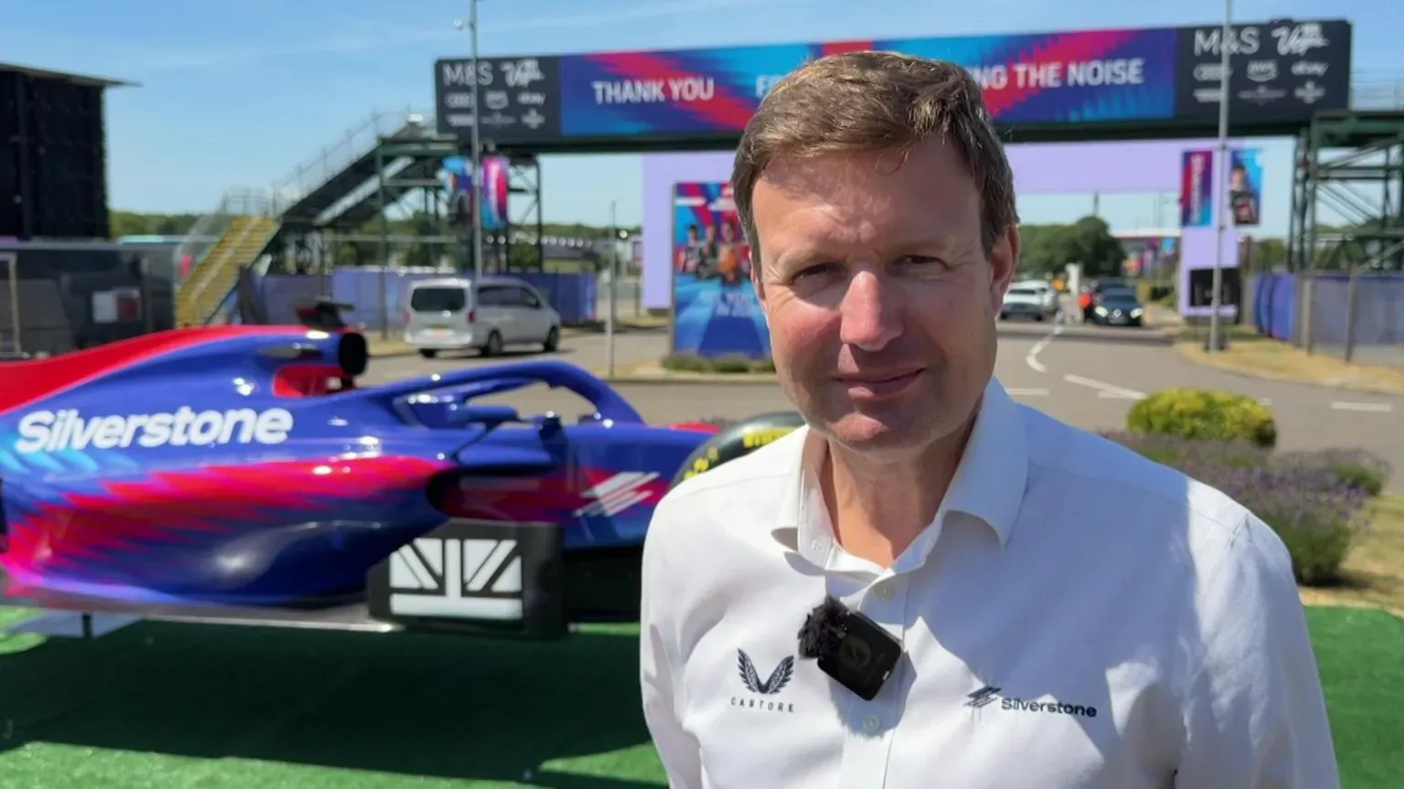 BBC A head and shoulders shot of Stuart Pringle, outside at Silverstone. He has short, light brown hair and is wearing a white open-neck shirt, with a small black microphone attached to it. Behind him is a purple and red car with Silverstone written on it in white letters and beyond that a road with a bridge over it.