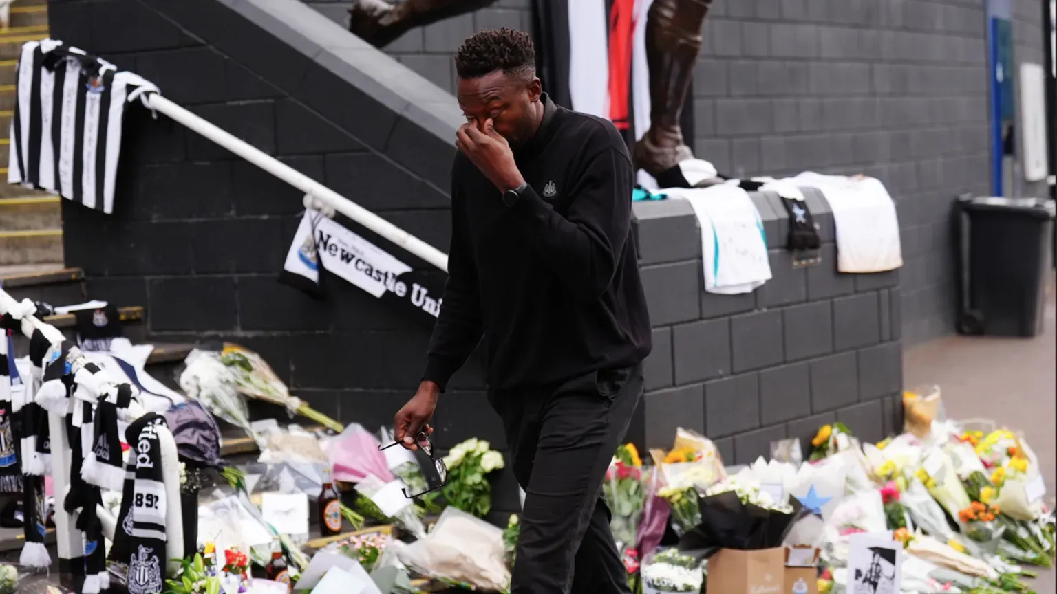 PA Media Shola Ameobi, dressed in black, wipes away tears as he leaves St James' Park. The steps behind him are covered in flowers with Newcastle United shirts and scarves hanging from the banisters.